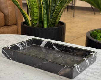 May include: A rectangular black marble tray with white veining. The tray has a raised edge and a smooth interior surface. The tray is sitting on a white marble surface. The background includes potted plants.