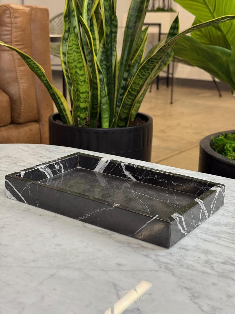 May include: A rectangular black marble tray with white veining. The tray has a raised edge and a smooth interior surface. The tray is sitting on a white marble surface. The background includes potted plants.