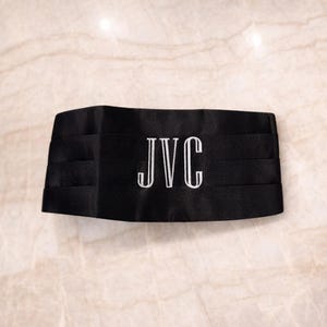 Puede incluir: Fajín negro con iniciales blancas bordadas "JVC". El fajín está hecho de tela plisada y está diseñado para ser usado con ropa formal. Las iniciales están centradas en la parte delantera.