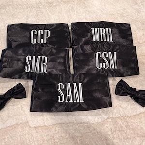 May include: Black satin cummerbunds and bow ties. Each cummerbund is embroidered with white initials. The initials are CCP, WRH, CSM, SMR, and SAM. The bow ties are also black satin.