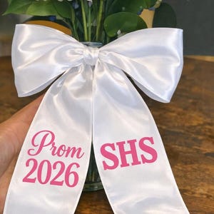 Könnte beinhalten: Eine weiße Satinschleife mit rosa Text „Prom 2026 SHS“ ist um eine Vase mit weißen und blauen Blumen gebunden. Die Enden der Schleife sind sichtbar, mit dem Text darauf gedruckt. Die Schleife ist der Mittelpunkt.