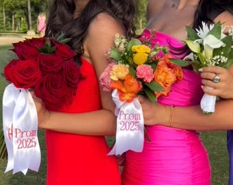 Puede incluir: Dos ramos de flores, uno con rosas rojas oscuras y otro con una mezcla de flores coloridas, son sostenidos por personas que visten vestidos brillantes. Cintas blancas con "Prom 2025" están unidas a los ramos.