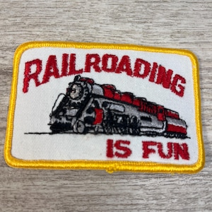 Può includere: Toppa ricamata con sfondo bianco, bordo giallo e le parole "RAILROADING IS FUN" in rosso. Un'illustrazione dettagliata di un treno è presente al centro, con accenti rossi e argento.