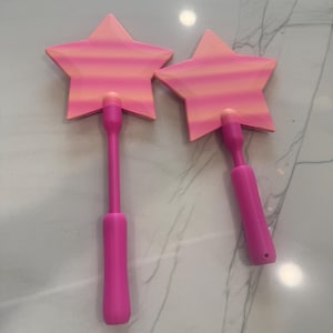 May include: Two pink star-shaped wands with a gradient of orange and pink. The wands have a long, thin handle and a rounded base. The star-shaped top is a light pink with a gradient of orange and pink.
