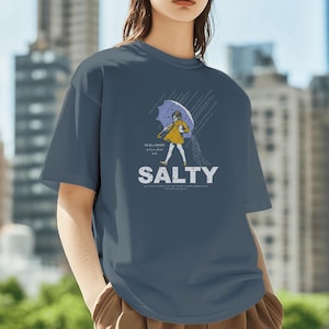 Op de afbeelding: Marineblauw t-shirt met een afbeelding van een persoon onder een paraplu, met het woord "SALTY" in witte letters. De persoon draagt een gele jas en houdt een zoutvaatje vast. Het shirt heeft korte mouwen en een ronde hals.