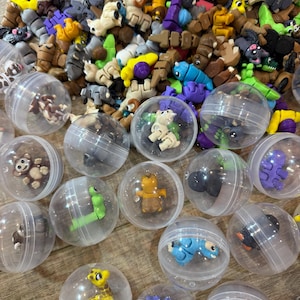 Op de afbeelding: Een verzameling kleine, kleurrijke dierenbeeldjes, waaronder beren, apen en pinguïns, wordt getoond. Sommige beeldjes zitten in doorzichtige plastic capsules. De beeldjes zijn in verschillende kleuren, waaronder bruin, blauw, geel en paars.