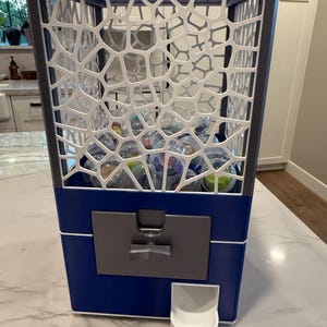 Puede incluir: Una máquina expendedora azul y blanca con un marco geométrico blanco similar a una red que rodea un área de almacenamiento transparente llena de pequeñas cápsulas de plástico redondas. La base es azul con un mecanismo gris y una salida.