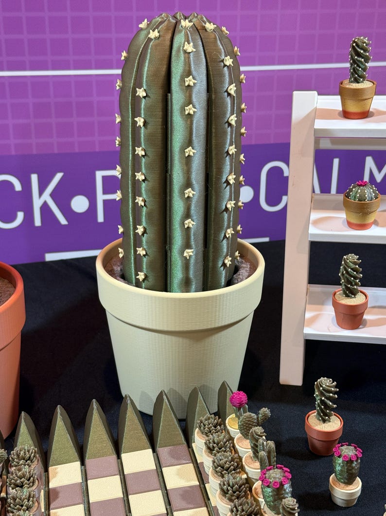 May include: A large, olive-green cactus in a beige pot, adorned with small, star-shaped accents. The image also features a white shelf with miniature cacti in terracotta pots, and a row of decorative cactus-shaped fence pieces.