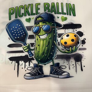 Op de afbeelding: Een cartoonillustratie van een augurk met een zonnebril en een baseballpet achterstevoren, met een pickleball racket en bal. De tekst "PICKLE BALLIN" staat boven de augurk. De augurk draagt hoge sneakers.
