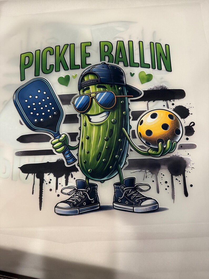 Op de afbeelding: Een cartoonillustratie van een augurk met een zonnebril en een baseballpet achterstevoren, met een pickleball racket en bal. De tekst "PICKLE BALLIN" staat boven de augurk. De augurk draagt hoge sneakers.