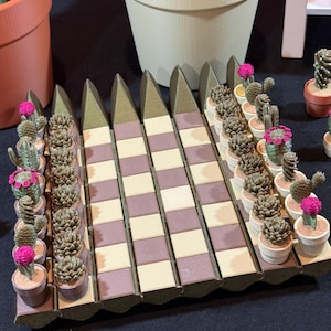 May include: A display of miniature cactus plants in small pots, arranged on a decorative, geometric platform. The cacti feature various shapes and are adorned with pink flowers. The platform has a brown and beige checkered pattern.