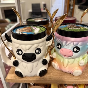 Op de afbeelding: Twee keramische mokken in de vorm van cartoonkoeien, één wit en één pastelkleurig, elk met een gouden lepel. De mokken staan op een houten plank, met de deksels van de ijscontainers zichtbaar. De witte koeienmok heeft zwarte accenten.