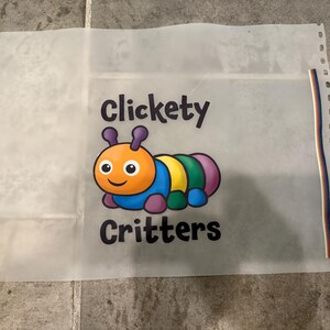 Op de afbeelding: Een doorschijnend vel met de woorden "Clickety Critters" en een kleurrijke cartoonrups. De rups heeft een oranje kop, een blauw lichaam en segmenten van geel, groen en paars. Het vel heeft gaten langs de linkerrand.