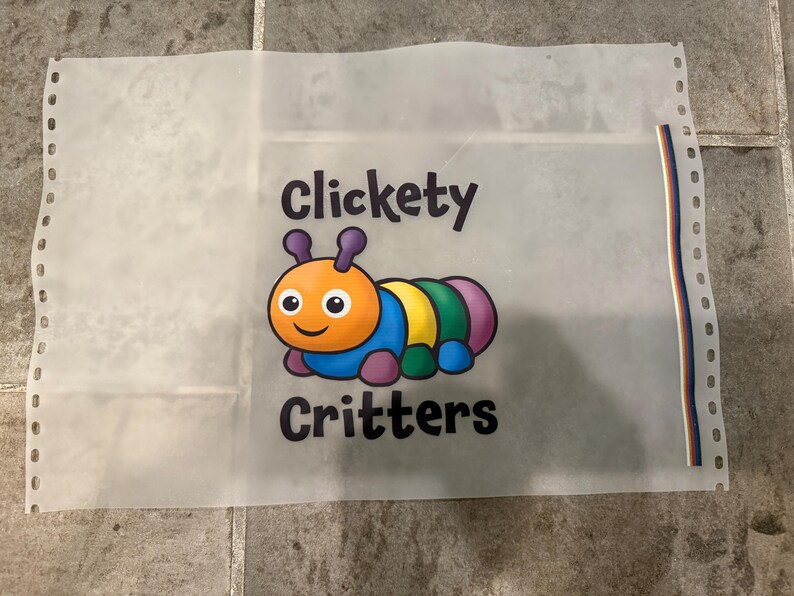 Op de afbeelding: Een doorschijnend vel met de woorden "Clickety Critters" en een kleurrijke cartoonrups. De rups heeft een oranje kop, een blauw lichaam en segmenten van geel, groen en paars. Het vel heeft gaten langs de linkerrand.