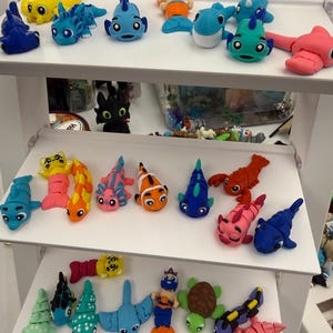 May include: A display of colorful, handmade clay figurines shaped like various sea creatures, including fish, a turtle, and a manta ray. The figurines are arranged on white shelves, showcasing a variety of colors and designs.