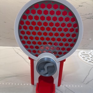 Puede incluir: Una máquina de chicles roja, blanca y gris con un diseño circular. La máquina presenta un patrón de panal de abeja rojo detrás de una rejilla circular gris. La base y el área de dispensación son rojas.