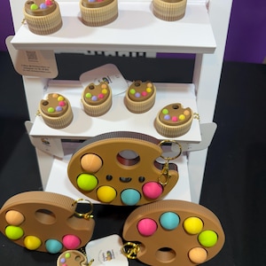 May include: A display of brown and tan keychains and toys. The keychains are shaped like paint palettes and cupcakes, with colorful, round accents. The toys have multiple colored bubbles. The display is on a white shelf.