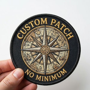 Op de afbeelding: Ronde, zwarte geborduurde patch met gouden tekst en een gedetailleerd kompasroosontwerp. De tekst luidt "CUSTOM PATCH" en "NO MINIMUM". De kompasroos heeft een kaartachtergrond.