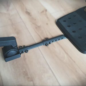 Può includere: Set di pedali da gioco neri con il logo Logitech G su una base nera. Il set è collegato a un braccio regolabile nero con più fori per viti. L'installazione è su un pavimento in legno marrone chiaro.