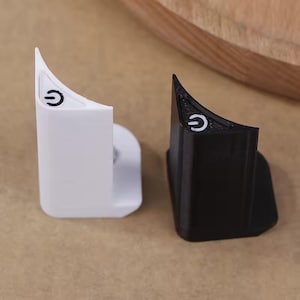 Może przedstawiać: Dwa plastikowe uchwyty w kształcie przycisku zasilania, jeden biały i jeden czarny. Każdy ma zakrzywioną górę z czarnym symbolem zasilania. Uchwyty są przeznaczone do trzymania czegoś, prawdopodobnie urządzenia lub akcesorium.