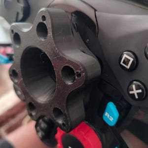 Può includere: Primo piano di un controller di gioco nero con un accessorio nero personalizzato. Il controller presenta pulsanti blu e rossi e un D-pad nero. L'accessorio ha più fori e un design circolare.