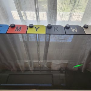 Op de afbeelding: Een set van zes rechthoekige inktcartridge-tabs op een rij, elk voorzien van een zwarte ronde knop aan de bovenkant. De tabs zijn blauw, rood, geel, zwart, donkergrijs en wit van kleur en gelabeld met de letters C, M, Y, K, W en G.