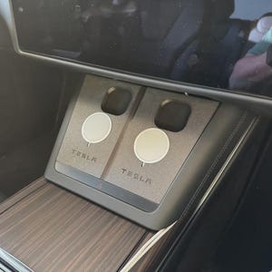 Peut inclure: Intérieur de voiture avec une console centrale comprenant un socle de chargement sans fil pour deux appareils. Le socle est gris et porte l'inscription "TESLA". La console est également ornée d'une garniture en bois.
