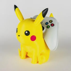 Könnte beinhalten: Ein gelber Pikachu-förmiger Gaming-Controller-Halter mit schwarzspitzigen Ohren und einer roten Wange. Ein weißer und grauer Gaming-Controller ist in den Halter eingesetzt. Der Controller hat farbige Tasten.