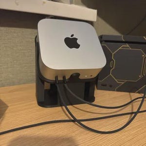 May include: A silver Apple Mac Mini computer with the Apple logo on top, mounted in a black stand. Several black cables are connected to the computer. The computer sits on a wooden surface.