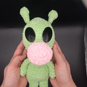 Peut inclure: Une peluche crochetée vert clair dotée de deux antennes dressées, de grands yeux ovales en plastique noir et d'un museau rond en crochet rose. La texture douce du fil donne à la créature un corps arrondi et de petits membres.