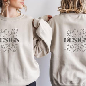 Może przedstawiać: Dwie beżowe bluzy z nadrukiem "YOUR DESIGN HERE" z przodu i z tyłu. Bluzy są w neutralnym kolorze, a tekst jest w stylowej czcionce. Mockup do projektowania odzieży.