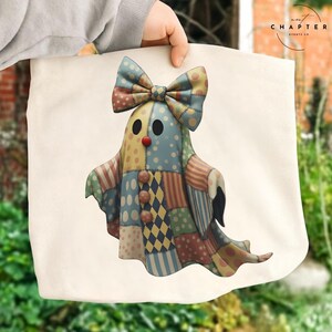 May include: A cream-colored tote bag featuring a whimsical patchwork ghost design. The ghost has a large bow, a red nose, and is made of various patterned fabrics in shades of blue, yellow, and red. The bag is held by a person.
