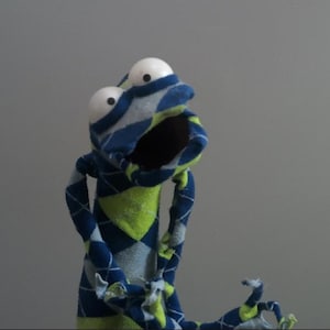 Op de afbeelding: Een handpop gemaakt van stof met een argyle-patroon in blauw, lichtblauw en limoengroen. De pop heeft twee grote, ronde, witte ogen met zwarte pupillen en een open mond, met gerafelde stoffen randen bij de aanzet van de armen.