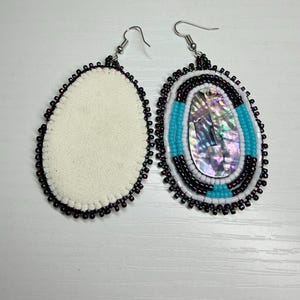May include: Oval-shaped beaded earrings with a white backing. One earring features a central iridescent stone, bordered by white, turquoise, and black beads. The other earring has a plain white backing, both with black beaded edges.