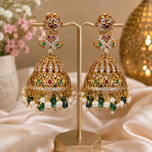 May include: A pair of gold-toned jhumka earrings featuring intricate filigree metalwork, embedded with clear white stones, small red and green accents, and dangling white pearls and green teardrop-shaped beads suspended from the bottom edge.