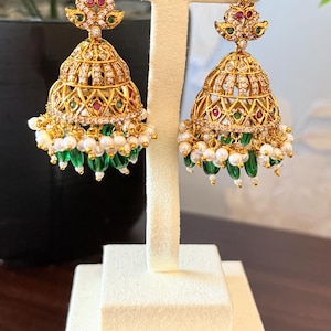 May include: A pair of gold-toned jhumka earrings featuring a bell-shaped design adorned with clear crystals, small red and green stones, and a dangling fringe of white pearls and green teardrop-shaped beads, suspended from a floral-shaped stud.