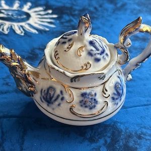 May include: A white porcelain teapot featuring blue floral patterns and gold trim accents on the spout, handle, lid, and body. The teapot has a scalloped rim and a decorative lid with a pointed finial, showcasing intricate gold detailing.