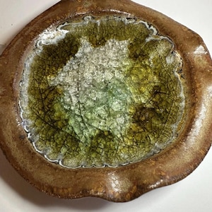 May include: A round ceramic dish with a wide, textured brown rim and a center filled with cracked, translucent green and clear glass. The interior surface features a distinct network of dark, fine lines creating a web-like pattern across the glass.