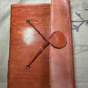 May include: A rectangular brown leather journal featuring a smooth finish and a wrap-around closure. The cover is secured by a thin red cord that loops through two metal eyelets and fastens around a circular leather disc attached to the side flap.
