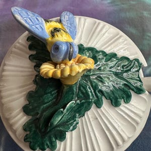 May include: A white ceramic lid featuring a three-dimensional sculpted bee with blue wings and a yellow body, resting on a yellow flower surrounded by dark green textured leaves. The lid has a radial ribbed pattern extending from the center.