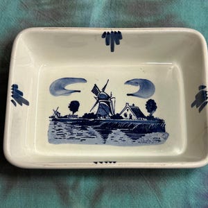 May include: A rectangular white ceramic dish featuring a blue hand-painted illustration of a Dutch windmill, a small house, and trees near a body of water. The rim is decorated with blue brushstroke accents at the center of each side.