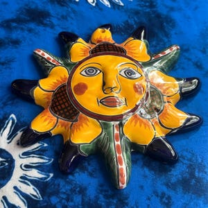 May include: A ceramic sun wall plaque featuring a central yellow face with black facial features, surrounded by yellow sunflower petals and dark blue pointed rays. The piece has a glossy finish and includes a small hole at the top for hanging.