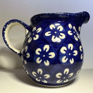 May include: A ceramic pitcher featuring a deep blue glaze decorated with a repeating pattern of white five-petaled flowers. The vessel has a rounded body, a curved handle, and a small pouring spout at the top rim, showcasing a glossy finish.