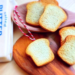 May include: A close-up shot of several toast-shaped ornaments with a light brown crust and a pale interior, arranged on wooden spoons and a wooden surface. A stick of salted butter is visible in the background.