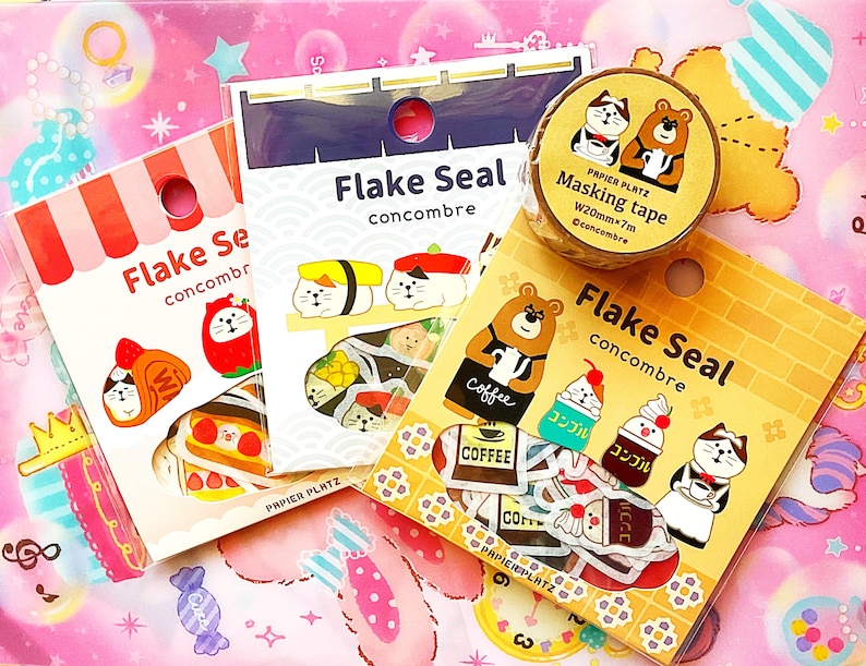 May include: An assortment of Japanese stationery items, including sticker packs and masking tape. The sticker packs feature cute illustrations of cats, bears, and food items. The masking tape roll has a cat and bear design. The text "Flake Seal concombre" is visible.