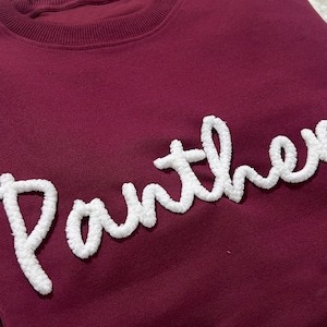 Può includere: Una felpa bordeaux con la scritta "Panther" ricamata in bianco, con lettere cursive e soffici. La felpa ha un girocollo e un design semplice, ideale per un uso casual.