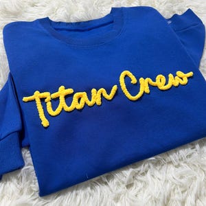 May include: A royal blue sweatshirt featuring the text "Titan Crew" in yellow, script-style lettering. The sweatshirt has a crew neck and long sleeves. The text appears to be made of a textured material.