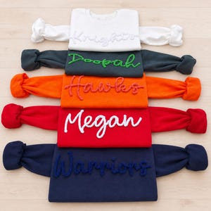 May include: Five long-sleeve sweatshirts stacked vertically in white, dark gray, orange, red, and navy blue. Each sweatshirt features a unique word embroidered in a textured, raised script font: Knights, Doopah, Hawks, Megan, and Warriors.