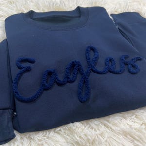 May include: A navy blue sweatshirt featuring the word "Eagles" in a textured, darker blue embroidery. The crew neck sweatshirt has long sleeves. The item is presented on a white, fluffy surface.