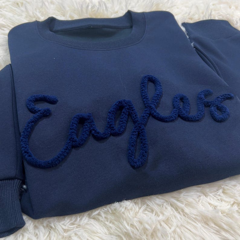 May include: A navy blue sweatshirt featuring the word "Eagles" in a textured, darker blue embroidery. The crew neck sweatshirt has long sleeves. The item is presented on a white, fluffy surface.